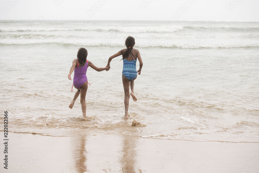 Rear view of two mixed-race sisters running into the water playing and laughing having fun on the beach on a bright tropical summer holiday