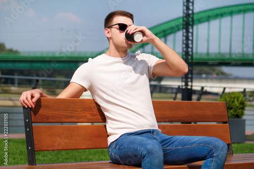 Handsome boy wearing sunglasses, sitting on a bench on a pier and drinking coffee to go. Taking a break from city rush. Background scenery bridge, river, pier, path along the river and green grass.