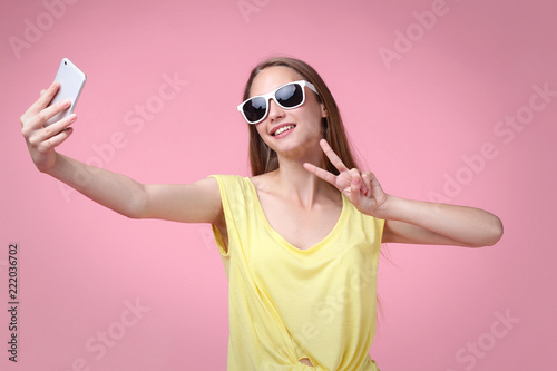Young woman in sunglasses making selfie on pink background