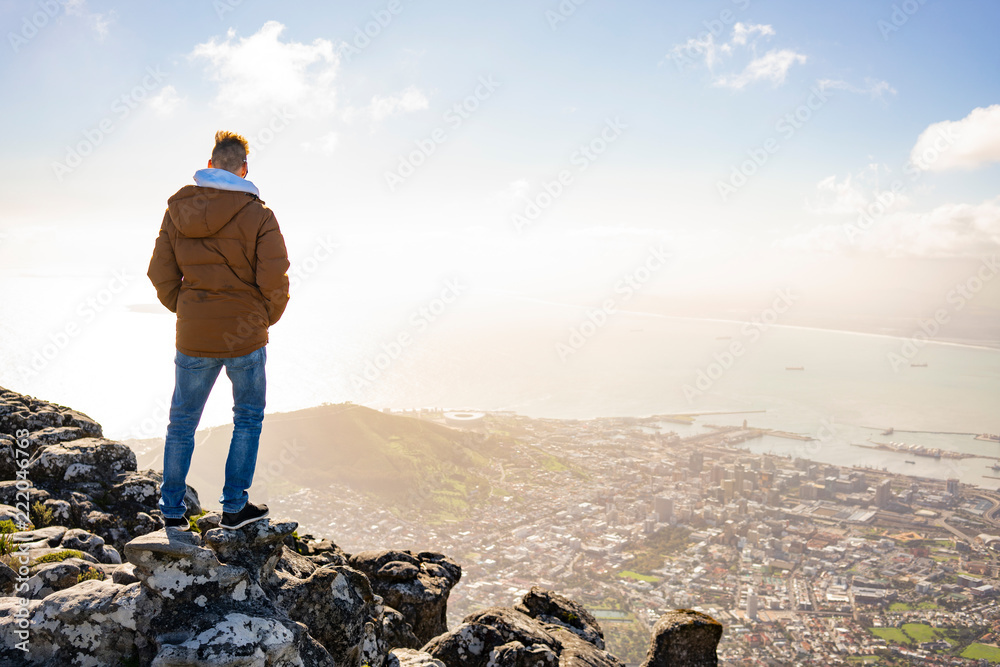 Fototapeta premium Young man is sitting on rock at Table Mountain and looking at Cape Town, South Africa