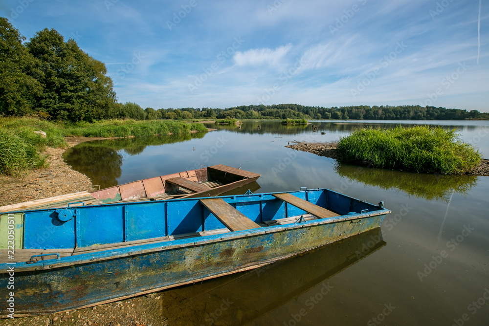 Blue and brown metal rowing boats for fishing chained on sandy lake shore, calm water surface, reflection in water, small green grass isle, sunny summer day, blue sky, white clouds