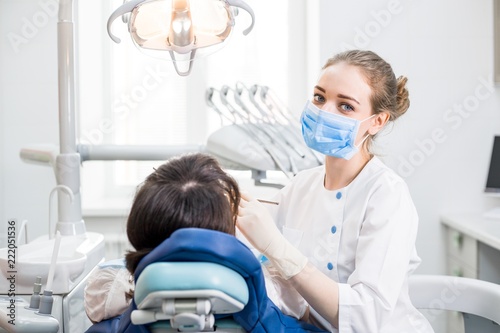 Beautiful girl dentist with blue eyes in white uniform examines the oral cavity of the patient