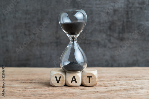 Time countdown for VAT increase deadline concept, hourglass or sandglass on cube wooden block with alphabets VAT, Value Added Tax on wood table with dark black loft cement wall photo