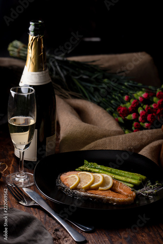 Fototapeta Naklejka Na Ścianę i Meble -  salmon steak with green asparagus and topping with sliced yellow lemon on black plate on woodden table with an empty glass of wine
