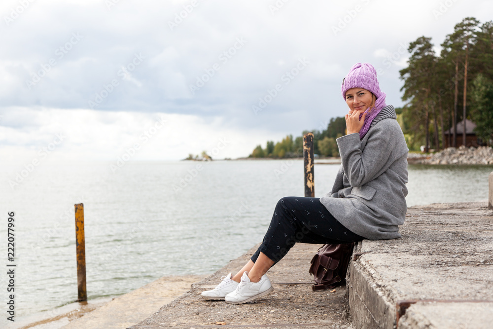 Outdoor atmospheric lifestyle photo of young beautiful  darkhaired woman  in knitting hat, in a gray coat and black trousers sits on the pier next to the sea  in sunny autumn day