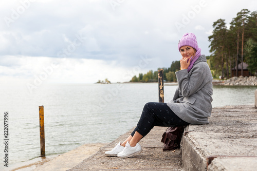 Outdoor atmospheric lifestyle photo of young beautiful darkhaired woman in knitting hat, in a gray coat and black trousers sits on the pier next to the sea in sunny autumn day