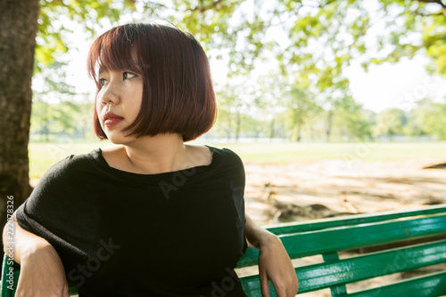 Young Asian woman sitting alone on the public bench in the park surrounded with nature and warm sunlight. Young lonely woman sitting alone in the park. Outdoor activity in the park concept.