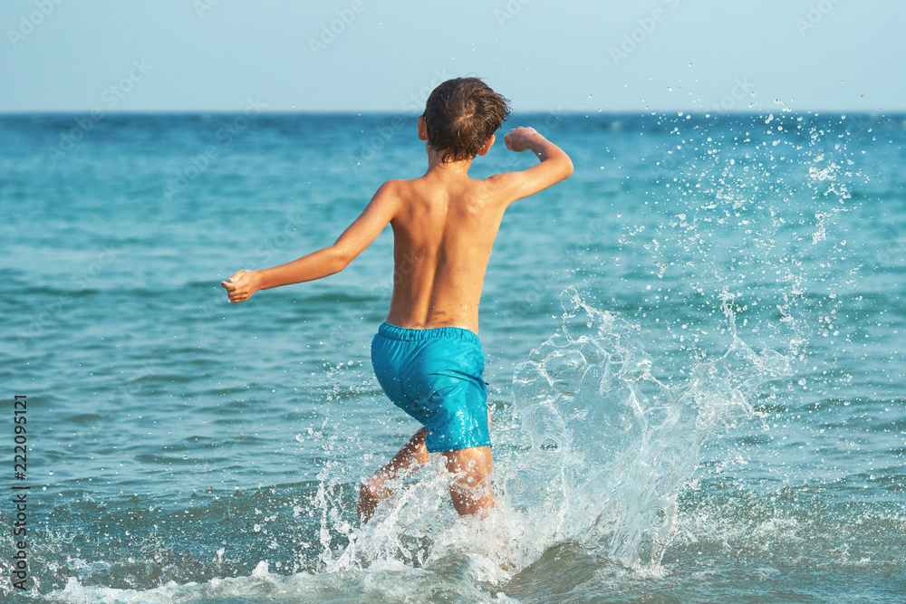 A happy little boy is happily running and splashing in the sea water in summer on a warm, sunny day. Large splashes of water close-up. A child is playing on the seashore A happy little boy is happily running and splashing in the sea water in summer on a warm, sunny day. Large splashes of water close-up. A child is playing on the seashore