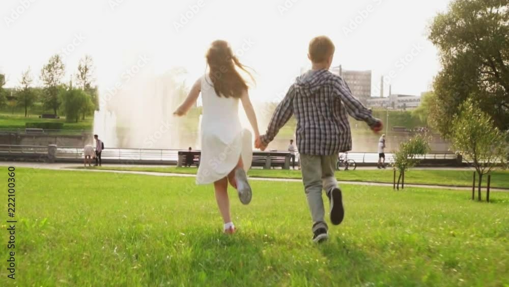 Carefree children holding hands and running together on grass in summer park