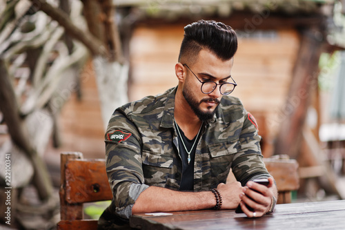 Awesome beautiful tall ararbian beard macho man in glasses and military jacket sitting outdoor wooden table of restaurant and looking his phone.