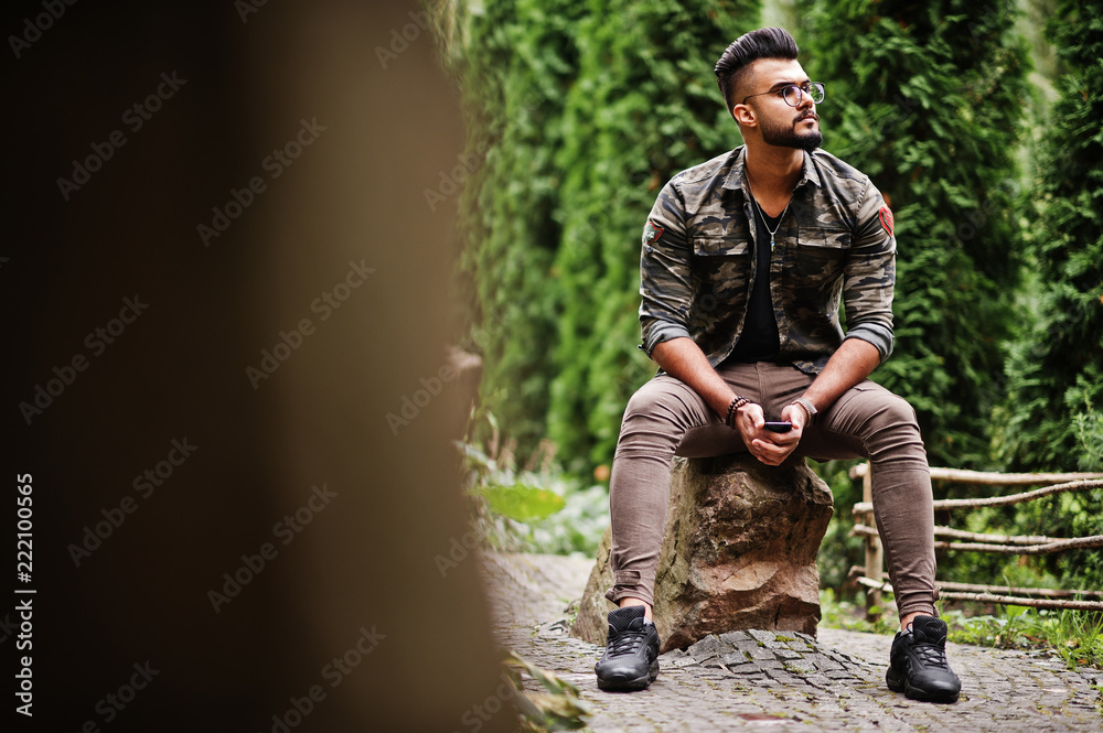 Awesome beautiful tall ararbian beard macho man in glasses and military jacket posed outdoor, sitting on stone.
