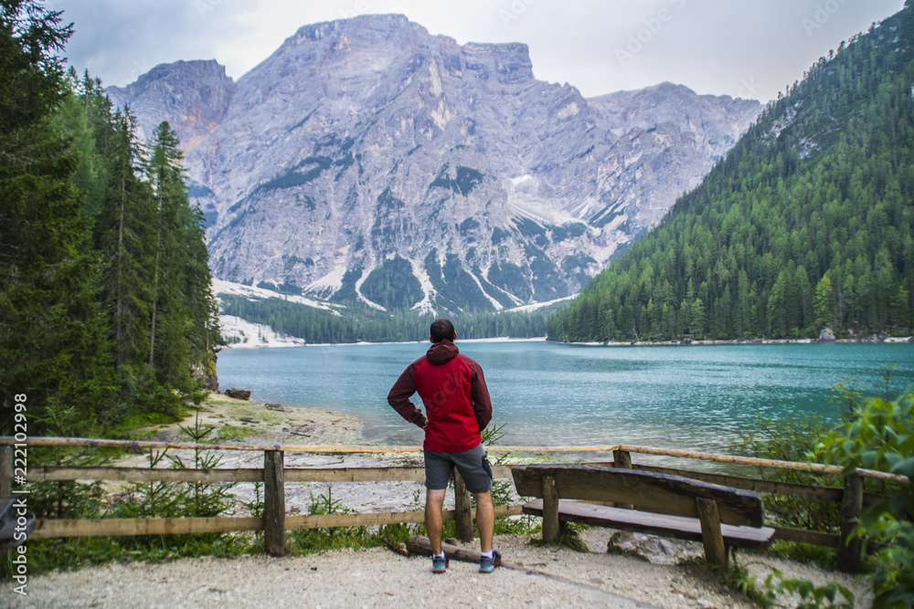 View of young tourist hiker man overlooking beautiful alpine lake Lago Di Braies (Pragser wildsee) in Trentino, Dolomites mountains, Italy. Tourist popular summer attraction/destination in Europe