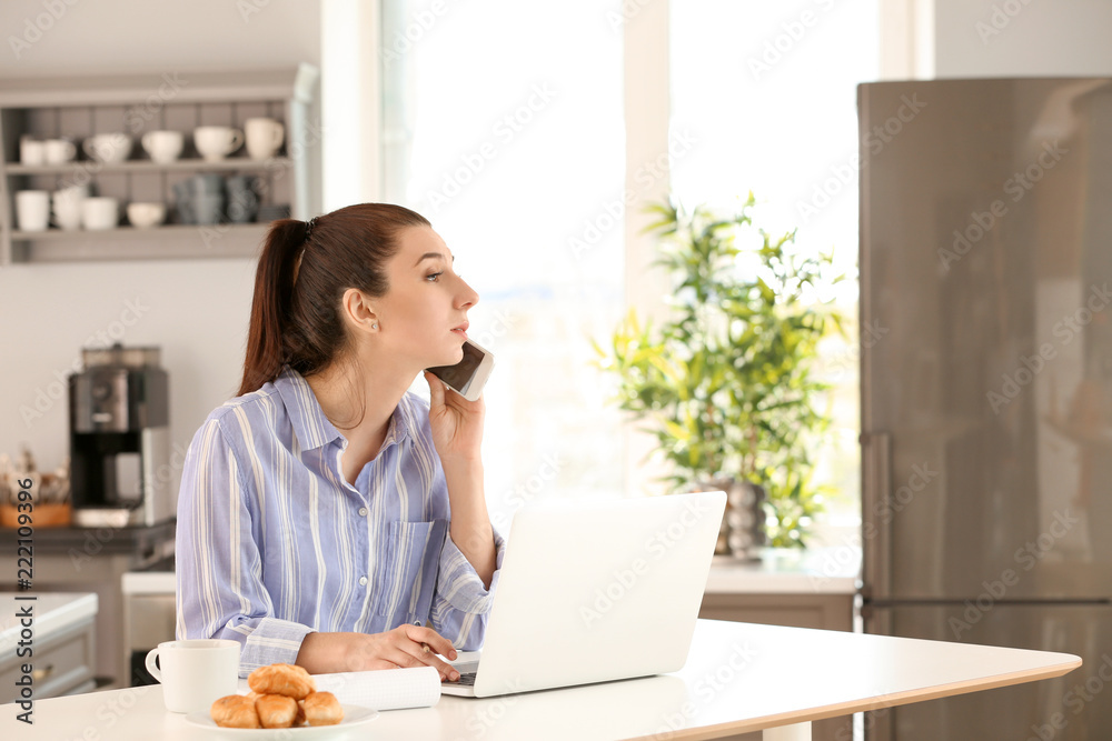 Young female freelancer talking on phone while working with laptop at home