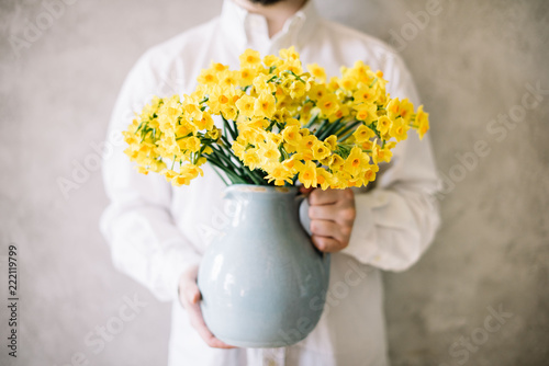 Fototapeta Naklejka Na Ścianę i Meble -  Very nice young man in a white shirt holding blossoming yellow Narcissus flower bouquet in a blue ceramic vase on the grey wall background