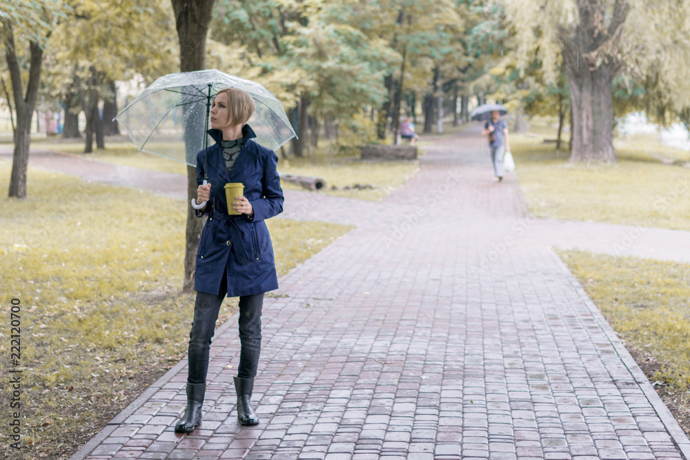 A woman of 30-40 years in a blue raincoat under a transparent umbrella walks in the park and drinks coffee.