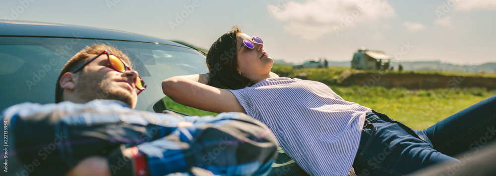 Young couple resting lying on the windshield and the hood of the car - obrazy, fototapety, plakaty 