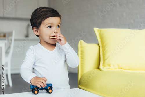 cute child playing with toy car and eating cookie at home