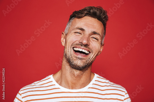Handsome guy with stubble in striped t-shirt laughing on camera, isolated over red background