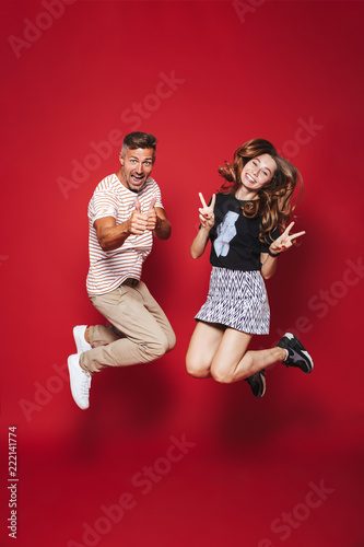 Full length photo of excited man and woman in striped t-shirt jumping, while showing victory sign, isolated over red background