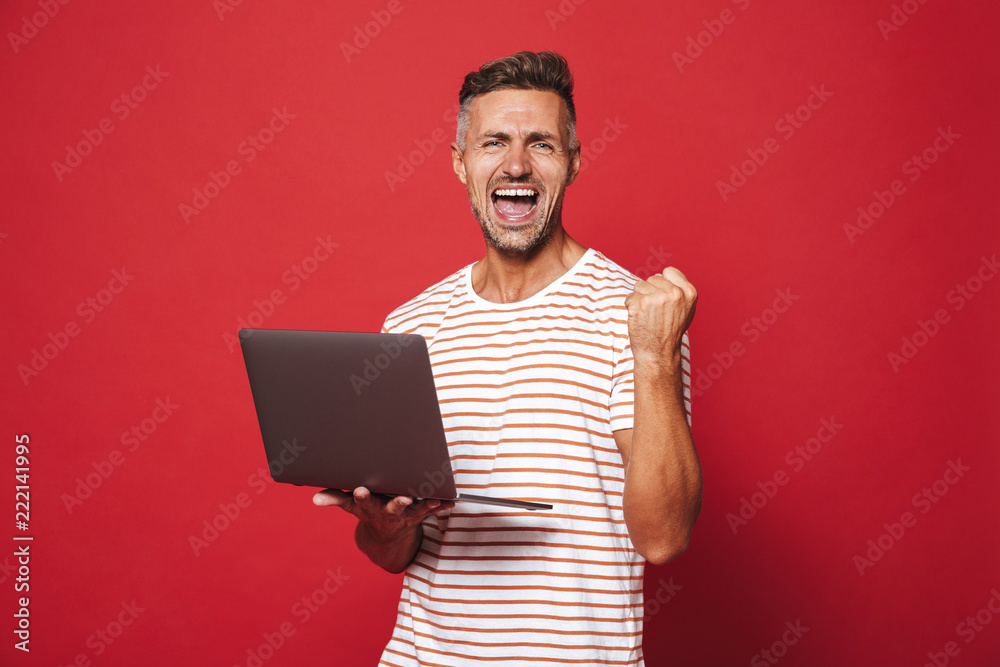 Image of successful guy in striped t-shirt smiling and holding gray laptop, isolated over red background Image of successful guy in striped t-shirt smiling and holding gray laptop, isolated over red background