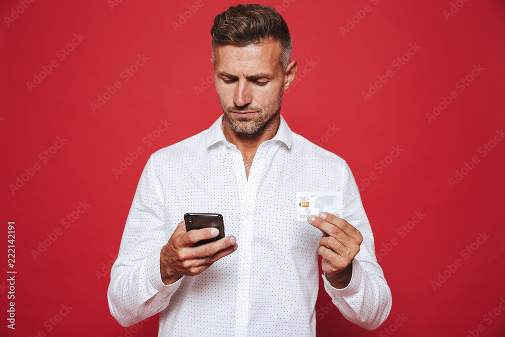 Photo of adult man in white shirt holding credit card and smartphone, isolated over red background Photo of adult man in white shirt holding credit card and smartphone, isolated over red background
