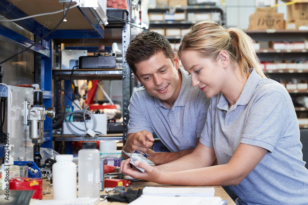 Engineer Helping Female Apprentice In Factory To Measure Component Using Micrometer Engineer Helping Female Apprentice In Factory To Measure Component Using Micrometer