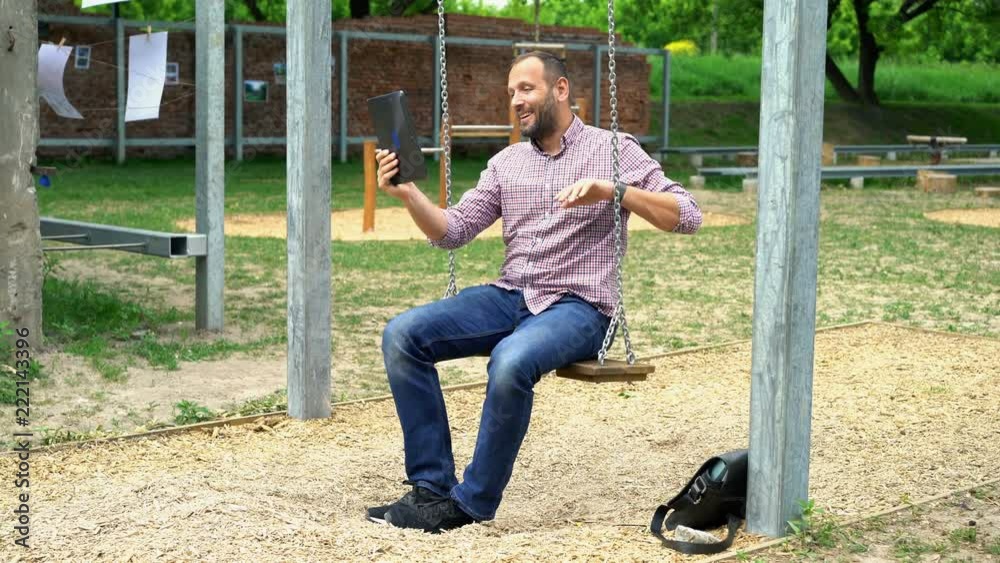 Young, happy man chatting on tablet sitting on swing in city park
Young, happy man chatting on tablet sitting on swing in city park