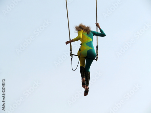 Girl aerial acrobat sitting on trapeze on sky background during a street circus performance. Woman gymnast performs acrobatic tricks