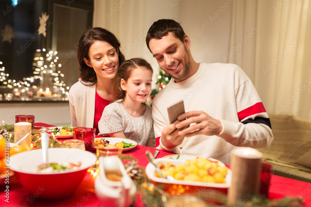 holidays, family and technology concept - happy mother, father and little daughter with smartphone having christmas dinner at home