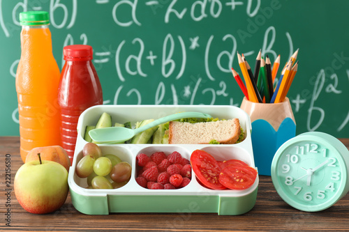 Fototapeta Naklejka Na Ścianę i Meble -  Lunch box with appetizing food and alarm clock on table near chalkboard