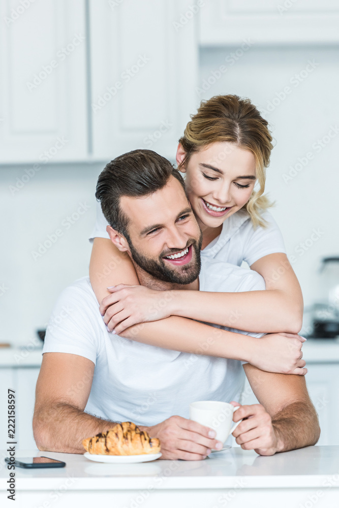 happy young woman hugging smiling boyfriend holding cup of coffee at morning