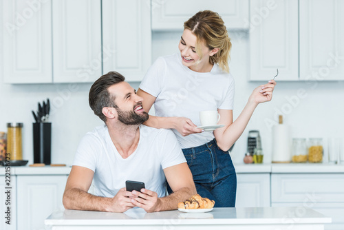 happy young man using smartphone and looking at smiling girlfriend drinking coffee at morning
