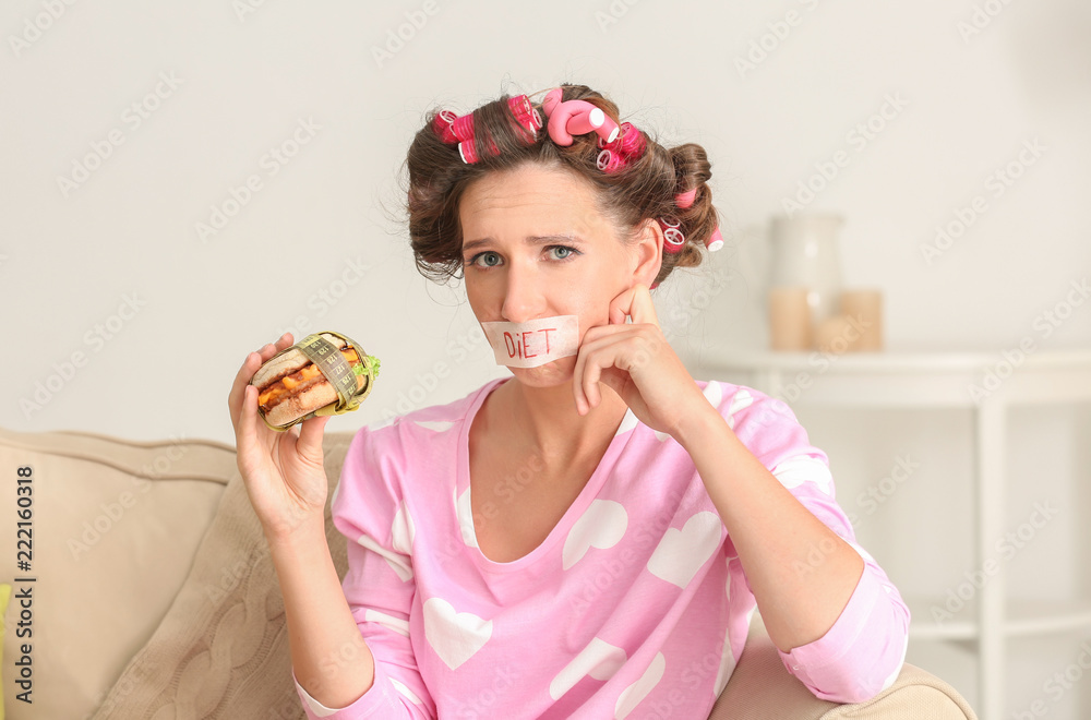 Emotional woman with taped mouth and tasty burger at home. Diet concept