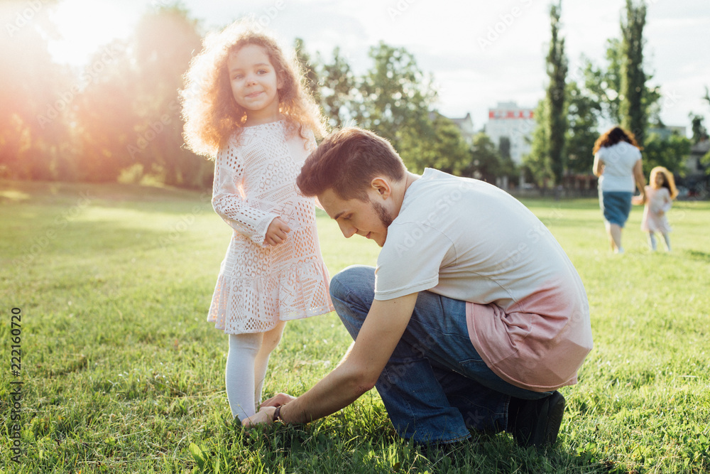 Fototapeta premium A father helping his little daughter with her shoes in the park. Cute curly kid girl in beautiful dress
