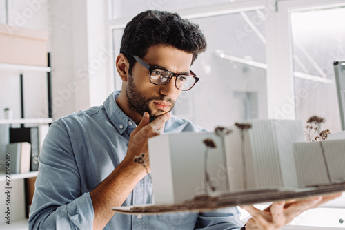 pensive architect in glasses touching chin and looking at architecture model in office