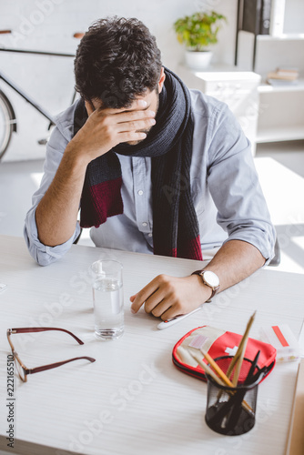 sick businessman in scarf touching forehead and sitting at table in office