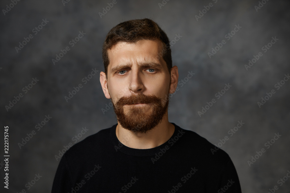 Isolated picture of handsome frustrated young male with thick beard and mustache frowning, biting lips and looking down with guilty facial expression, feeling ashamed and upset or being sorry