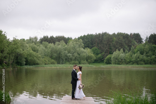Beautiful happy wedding couple of groom and bride standing on the pier near the lake and kissing. Wedding day concept