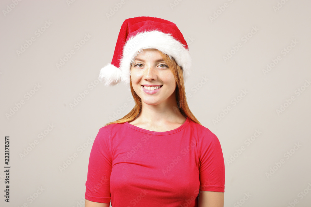 Close up portrait of beautiful redheaded young woman wearing Santa Claus hat and red t-shirt with satisfied facial expression. Cute female celebrating New Year. Copy space, isolated on gray background Close up portrait of beautiful redheaded young woman wearing Santa Claus hat and red t-shirt with satisfied facial expression. Cute female celebrating New Year. Copy space, isolated on gray background