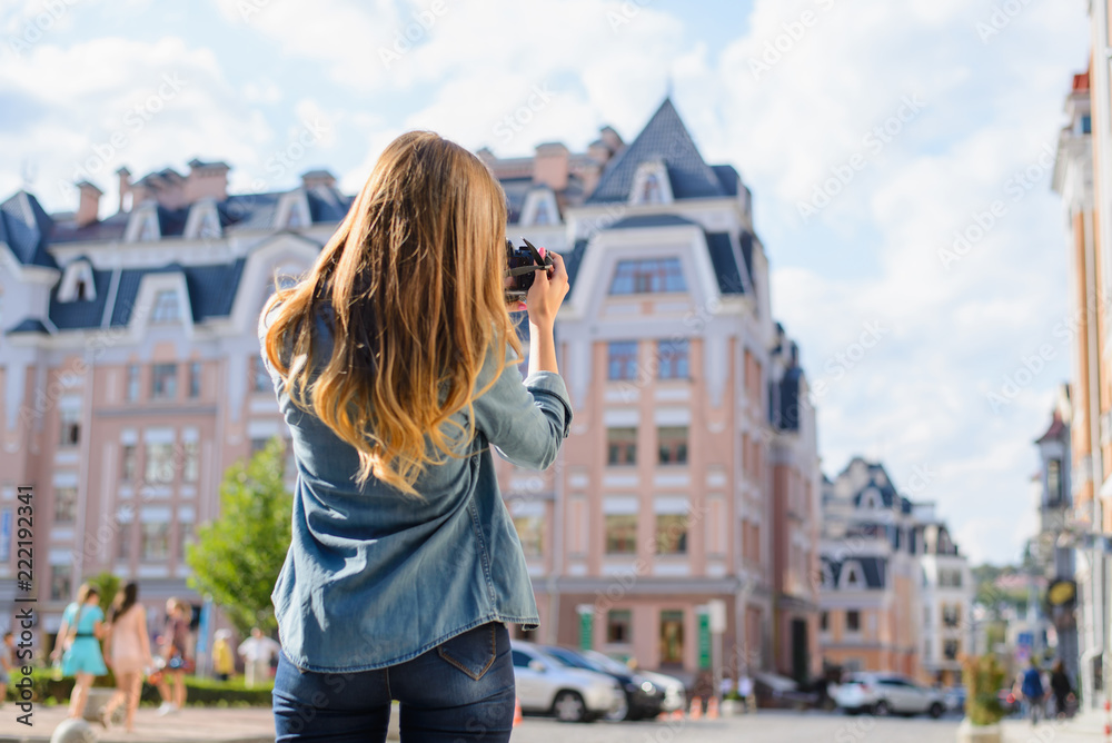 First day in new European city. Beutiful lady taking a photo of a house with digital camera. She is dressed in casual clothes. View from back, blured background