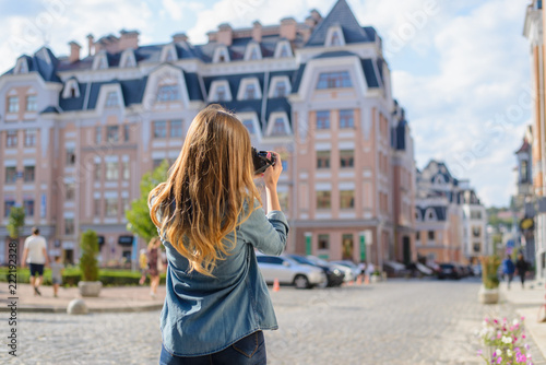 Happy, excited, stylish young woman taking photo of landmark in European city using her professional photo camera. She is dressed in jeand and shirt, she has lond blonde hair. View from back