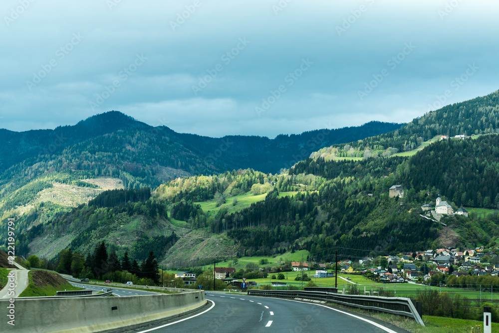 Autobahn und schmale Landstraße durch gebirgige Landschaft, bewölkt, teilweise sonnig, Dorf und Kirche auf einem Hügel