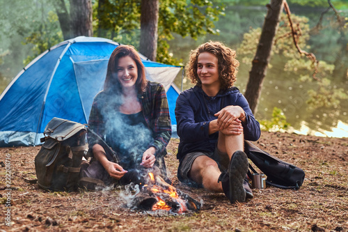 Young hiker couple sitting and talking while warming near a campfire at camp in the forest. Travel, tourism, and hike concept.