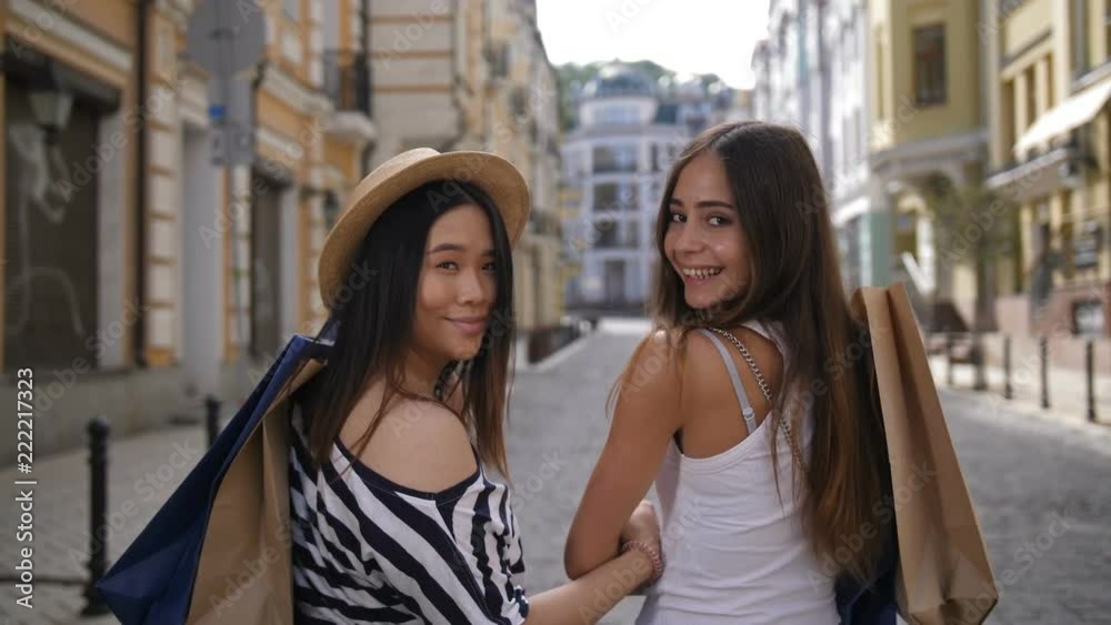 Beautiful young asian and caucasian female shopper friends walking with paper shopping bags on shoulders. Happy positive girls looking at camera and smiling with toothy smiles. Teenagers shopping Beautiful young asian and caucasian female shopper friends walking with paper shopping bags on shoulders. Happy positive girls looking at camera and smiling with toothy smiles. Teenagers shopping