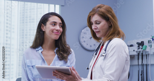 Portrait of beautiful young latina patient consulting with doctor about test results. Close up of pretty hispanic woman looking at tablet device with doctor indoors medical clinic