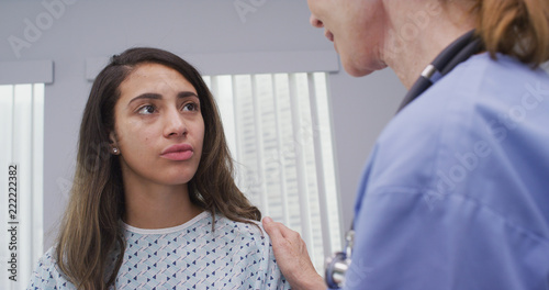 Medical nurse comforts young female patient holding her shoulder indoors hospital room. Mid aged nurse comforts young latina patient wearing hospital gown