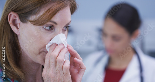 Attractive mid aaged patient blowing nose into tissue paper. Close up of caucasian woman wiping nose with tissue while doctor sits quietly in the background taking notes