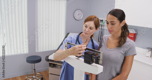 Mid aged caucasian nurse weighing female patient on scale. Charming middle aged nurse using scale to measure weight of young latina patient photo