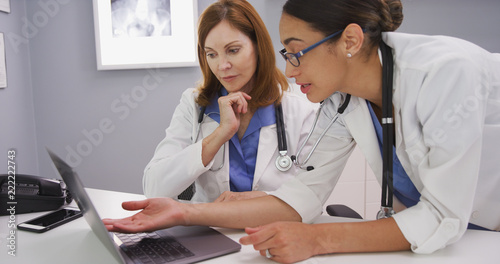 Intelligent young latina doctor looking at laptop computer and talking with elder physician. Two medical doctors using notebook computer to discuss patients healh condition photo