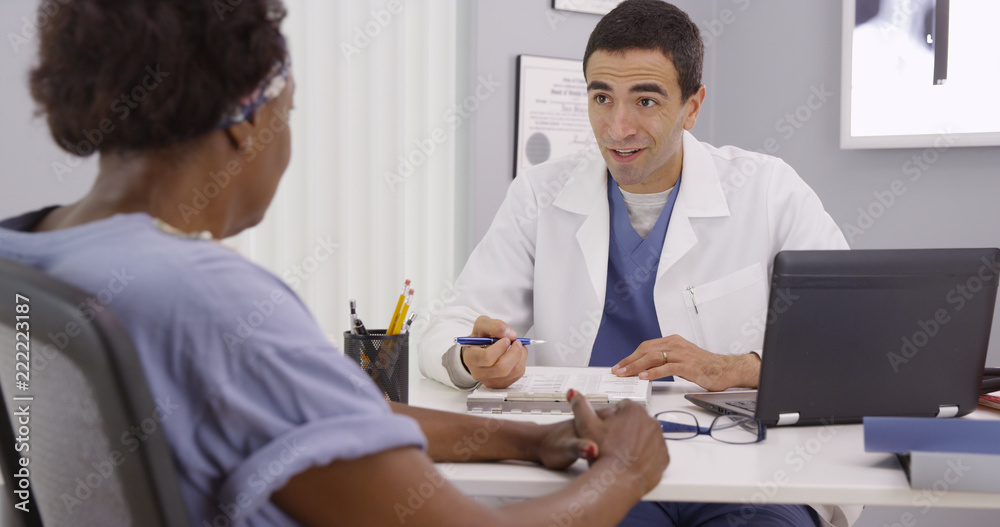 Young male doctor and mid aged patient sitting in medical office discussing health history. Senior black woman having appointment meeting with doctor about results from her mri Young male doctor and mid aged patient sitting in medical office discussing health history. Senior black woman having appointment meeting with doctor about results from her mri